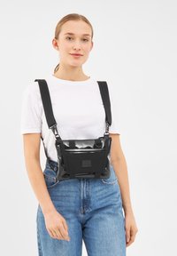 Borsa a tracolla nera e lucida con cinturini regolabili, dotata di una tasca frontale e di forma rettangolare. Indossata sopra una camicia bianca e jeans.