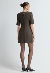 Hnědé krátké šaty s přiléhavým střihem, které mají zadní zip a výřez. Modelka nosí černé vysoké podpatky a jemné punčochy.