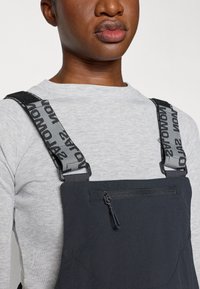 Črne džinsase s funkcionalnim dizajnom, sivo nastavljive naramnice z napisom "Salomon" in sprednjim žepom na zadrgo. Nosi se čez svetlo sivo majico.