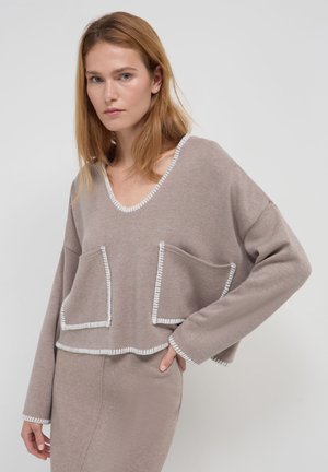 Maglione corto color marrone chiaro con scollatura a V, cuciture a contrasto bianche sui bordi e due tasche frontali. Tessuto morbido e vestibilità rilassata.