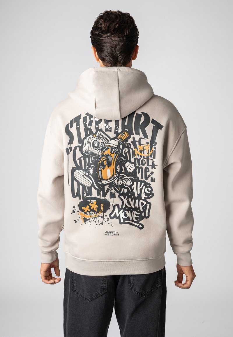 Beige capuchontrui met een graffiti-achtige print op de achterkant met gedurfde zwarte en oranje letters en illustraties. Zachte textuur.