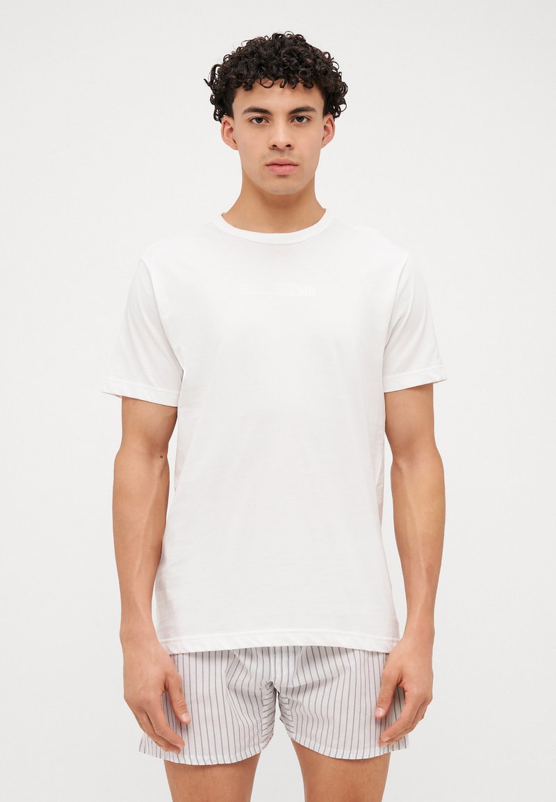 Camiseta blanca de algodón con mangas enrolladas y sutil logotipo de Calvin Klein. Combinada con pantalones cortos de rayas claras, mostrando un ajuste relajado.