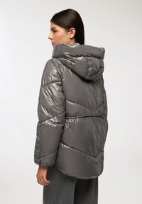 Giubbotto grigio imbottito con finitura lucida, cappuccio e cuciture a zigzag. Presenta una vestibilità rilassata e polsini elastici, progettato per calore e comfort.