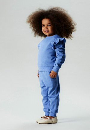 Jasnoniebieski dres z falbankami na rękawach, miękką fakturą i luźnym krojem, połączony z jasnym obuwiem. Prosty design, bez wzorów.