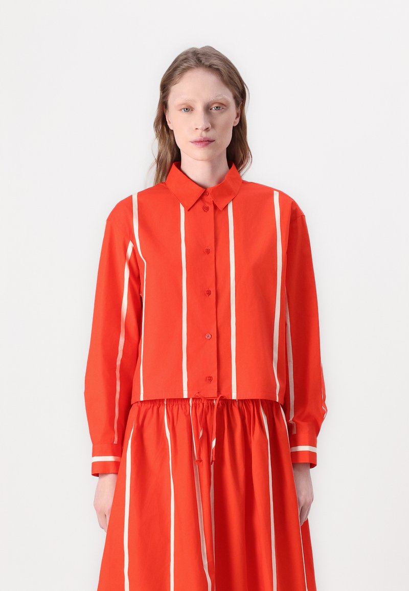 Camicia a righe rosse con colletto e maniche lunghe, caratterizzata da righe verticali bianche e chiusura a bottone. Il materiale appare leggero.