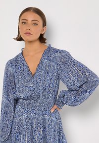 Blau gemustertes Kleid mit langen Ärmeln, tiefem V-Ausschnitt und gerafftem Taillenbereich. Es hat ein wirbelndes Blumenmuster in unterschiedlichen Blautönen.