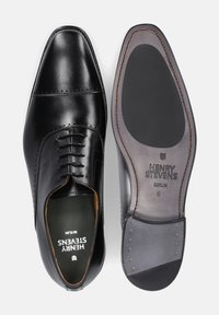 Schwarze Lederschuhe mit einem schlanken Design, spitzem Zehenbereich, Brogue-Details und einer braunen und schwarzen Sohle, die mit "HENRY STEVENS BERLIN" beschriftet ist.