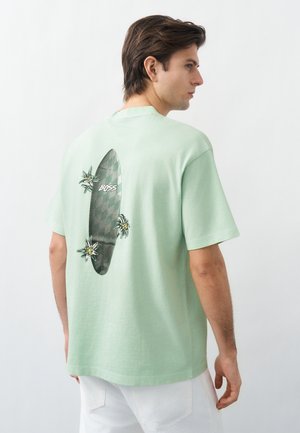 Νεαρός άνδρας που φοράει ανοιχτό πράσινο T-shirt με μαύρο σχέδιο σερφίστρας και λουλουδιών και τη λέξη "BOSS" στην πλάτη, συνδυασμένο με λευκό παντελόνι.