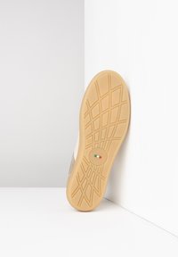 Semelle d'une basket avec une base en caoutchouc texturé, présentant un logo circulaire, inclinée contre un fond blanc.