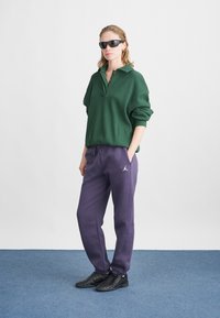 Polo de punto verde con cuello, combinado con pantalones sudadera morados con un logo, y zapatillas deportivas negras. El modelo lleva gafas de sol.