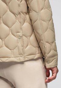 Giacca trapuntata beige con motivo a punto rotondo, bottoni a pressione ai polsini e sul davanti, indossata con pantaloni texturizzati beige chiaro.