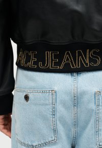 Μαύρο μπουφάν με κεντημένο χρυσό κείμενο "ACE JEANS" στο τελείωμα, συνδυασμένο με ανοιχτό μπλε τζιν παντελόνι που δείχνει τις πίσω τσέπες και λεπτομέρεια με κουμπί.