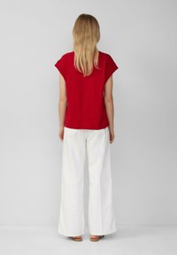 Vrouw met lang blond haar, gekleed in een rode top met korte mouwen, witte wijde broek en sandalen, staande tegen een effen grijze achtergrond met haar rug naar de camera gekeerd.