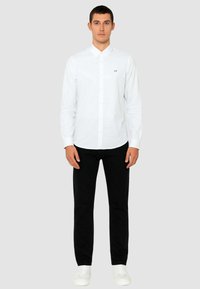 Camicia bianca a maniche lunghe con bottoni sul davanti e un piccolo logo "AX" sul petto, abbinata a pantaloni neri e scarpe bianche.
