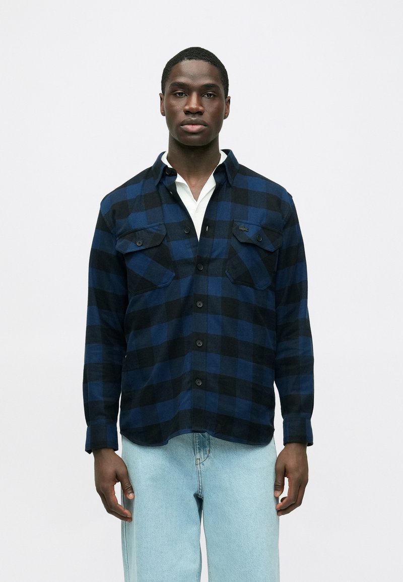 Camicia di flanella scozzese blu e nera con due tasche sul petto, bottoni frontali e colletto. Indossata sopra una maglietta bianca e jeans azzurri.