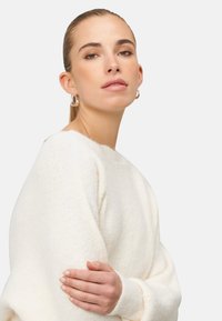 Sweter w kolorze kremowym o miękkim, teksturowanym wykończeniu i szerokim dekolcie. Modelka ma rękę na ramieniu i nosi srebrne kolczyki w kształcie kół.