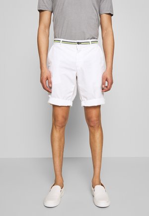 Man wearing rolled-up white shorts with striped belt, gray t-shirt, and white slip-on shoes standing against plain background.