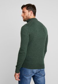 Suéter de punto verde oscuro con cuello alto, puños y dobladillo de canalé, diseño ajustado, combinado con jeans azules. Textura suave, estilo casual.