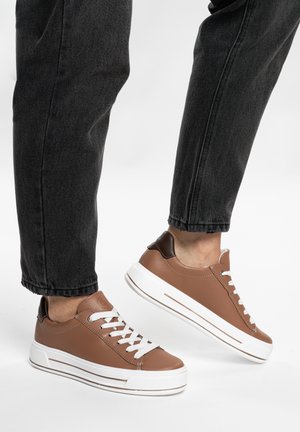 Person wearing brown leather sneakers with white soles and laces paired with black rolled-up jeans, showing one foot raised.