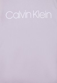 Camiseta de algodón de color púrpura claro con el nombre de la marca "Calvin Klein" en una fuente blanca grande en el pecho. Textura suave, diseño casual.