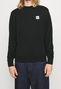 Osoba v černém sweatshirtu s dlouhým rukávem Lacoste a modrých džínách, s viditelným logem Lacoste na levé části hrudi.