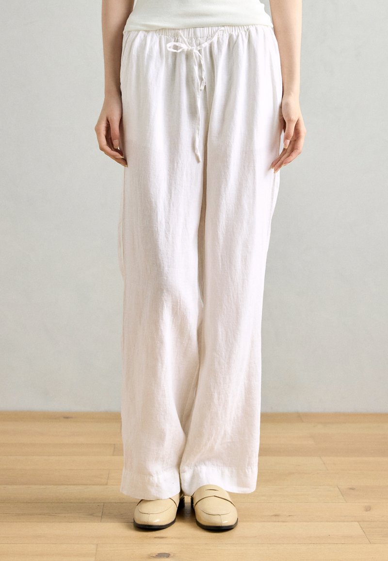 Pantalons larges en lin blanc avec une taille élastique et des détails de cordon de serrage. Texture lisse, coupe ample et longueur complète. Portés avec des chaussures beiges.