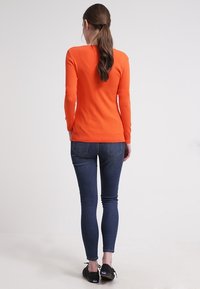 Maglione arancione brillante a maniche lunghe abbinato a jeans skinny blu scuro. Il maglione presenta un orlo a coste e un design aderente. Sneakers nere completano il look.