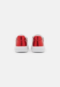 Accents arrière métalliques rouges sur des baskets blanches, avec une silhouette basse et une texture lisse, dotées d'une semelle en caoutchouc et d'un design minimaliste.