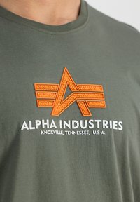 Zaļš kokvilnas T-krekls ar oranžu teksturētu logotipu ar spārniem un zīmola nosaukumu "ALPHA INDUSTRIES" baltā krāsā.