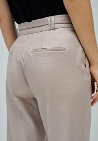 Persona con pantalones de tiro alto beige, con pasadores y bolsillos traseros con abertura, vestida con una blusa negra, con la mano descansando a un lado.