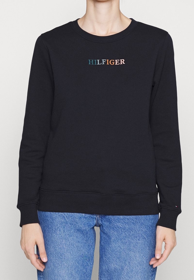 Personne portant un sweat-shirt noir à col rond avec le texte multicolore « HILFIGER » et un jean bleu, les bras détendus le long du corps.