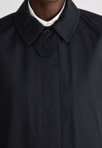 Abrigo negro con cuello puntiagudo, cierre de botones y textura suave y mate. Presenta costuras mínimas y un diseño streamlined.