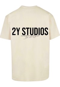 Kreminės spalvos marškinėliai su drąsiai juodu piešiniu ant nugaros: "2Y STUDIOS" su mažesniu tekstu apačioje "TIK BENDRUOMENĖS NARIAMS".