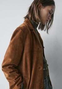 Bruine suède jas met een relaxte pasvorm, voorzien van een reverskraag, voorzakken en subtiele stiksel details, gedragen over een groene coltrui.
