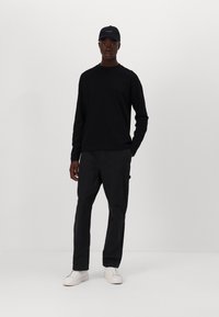 Schwarzes langärmliges T-Shirt, schwarze Cargo-Hosen und weiße Sneakers, ergänzt durch eine schwarze Mütze, vor einem schlichten weißen Hintergrund.
