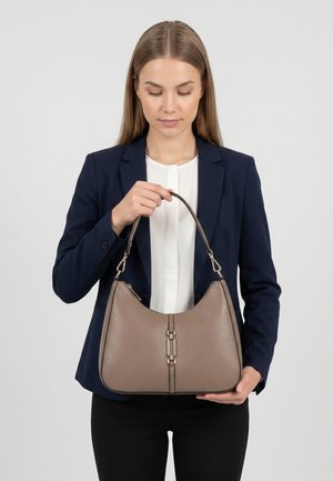 Donna in abbigliamento business che tiene una borsa in pelle taupe con chiusura metallica, guardando la borsa contro uno sfondo grigio uniforme.