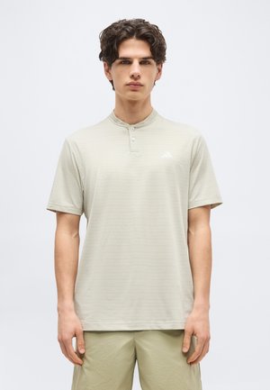 Jeune homme aux cheveux bouclés portant une chemise henley à manches courtes beige clair et un short assorti, sur un fond uni.