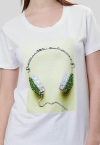 Camiseta blanca con un gráfico de auriculares. Los auriculares tienen lentejuelas verdes e iridiscentes, acentos plateados y un cordón decorativo.