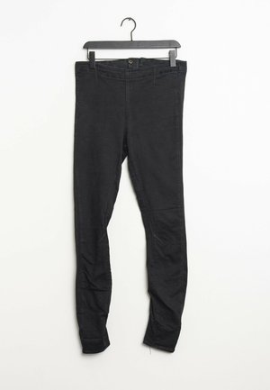 Pantaloni neri slim-fit appesi a una gruccia nera su uno sfondo bianco, che mostrano i dettagli dei bottoni frontali e delle cuciture.
