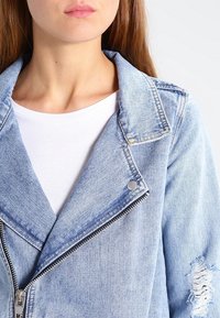 Světlá modrá džínová bunda s klopovým límcem, zipovým zapínáním a opotřebovanými detaily na rukávu. Nositá přes bílý tričko.