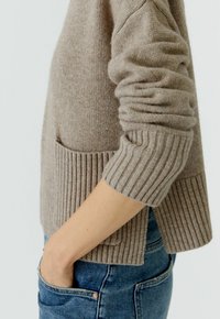 Beige gebreide trui met ribbels aan de mouwen en onderkant, met een zijsplit en een voork pocket. Draagt over blauwe jeans en toont een ontspannen pasvorm.