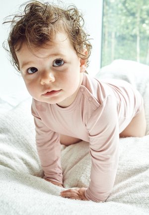 Lyserød langærmet bodystocking på en kravlende spæd med krøllet hår, som hviler på et struktureret hvidt tæppe. Blødt, glat stof tekstur synligt.