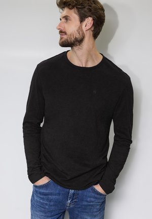 Camiseta negra de manga larga con cuello redondo y tela texturizada. Presenta un pequeño logotipo en el pecho, combinado con jeans de mezclilla azules.
