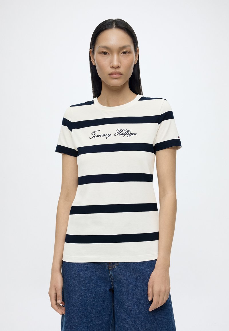 Γυναίκα με ίσια μαλλιά που φοράει λευκό T-shirt με μαύρες οριζόντιες ρίγες και το κείμενο «Tommy Hilfiger», συνδυασμένο με μπλε τζιν.