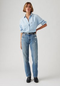 Chemise en denim bleu clair avec des manches retroussées, boutonnière à l'avant et poche poitrine, associée à un jean bleu déchiré taille haute et des bottines noires.