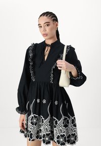 Μαύρο φόρεμα με λευκή floral κέντημα, με ρυάκι στον λαιμό και φουσκωτά μανίκια. Λεπτομερές δαντέλα στο τελείωμα; το μοντέλο κρατά μια μικρή λευκή τσάντα.