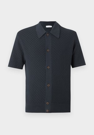 Homme aux cheveux bouclés foncés portant une chemise noire à manches courtes boutonnée, un jean bleu à jambes larges, des baskets blanches, debout une main dans la poche.