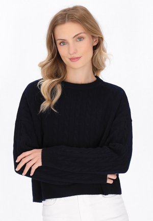Maglione bleu navy a lavorazione cable-knit con colletto rotondo a costine e maniche larghe, caratterizzato da motivi testurizzati, abbinato a jeans bianchi.