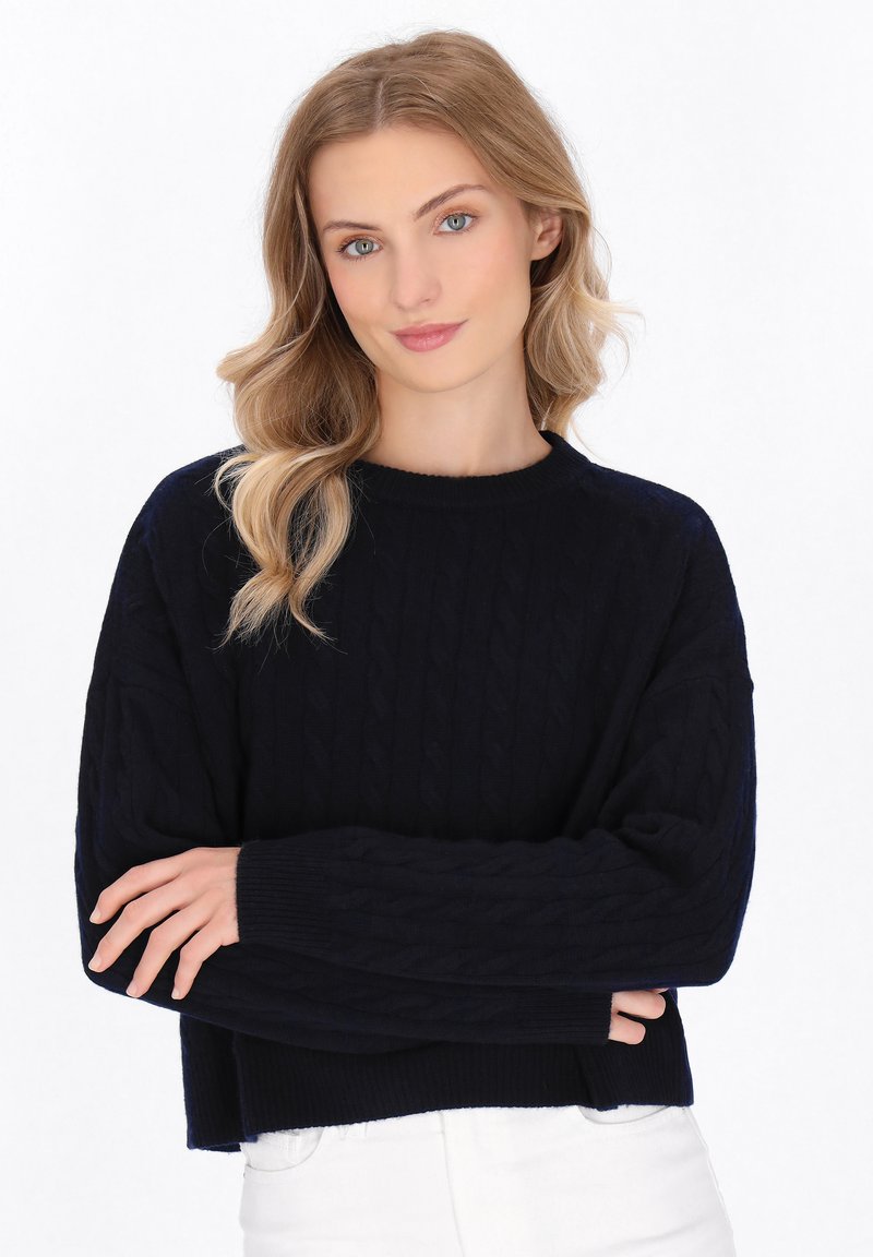 Maglione bleu navy a lavorazione cable-knit con colletto rotondo a costine e maniche larghe, caratterizzato da motivi testurizzati, abbinato a jeans bianchi.