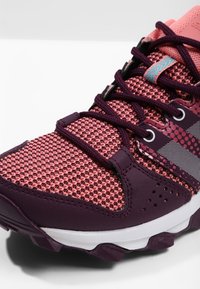 Zapatilla deportiva en púrpura y rosa con parte superior de malla, detalles texturizados y diseño estampado. Presenta cordones negros y una suela robusta.